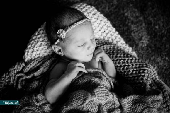 Artistiek zwart-wit portret van een pasgeboren baby die op de buik slaapt met de handjes onder de kin, rustend op een zachte ondergrond