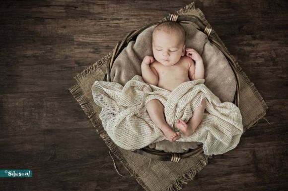Bovenaanzicht van een slapende pasgeboren baby, opgekruld in een zachte beige doek tegen een neutrale achtergrond.